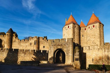 Ziyaretçiler Carcassonne 'un antik kale duvarlarının yanında yürürler. Günbatımının gölgeleri taş yapılar ve kulelerin üzerine düşüyor..