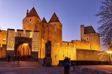 Ziyaretçiler, Fransa 'da gece çökerken sıcak ışıklardan parlayan Carcassonne' un antik duvarlarının yanında yürürler. Tarihi mekan pek çok kişiyi keşfetmeye çağırıyor.