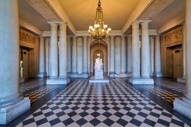 Ziyaretçiler Versailles Sarayı 'nın koridorunda yürüyorlar. Boşlukta sütunlar ve büyük bir avize var. Merkezde bir heykel sergileniyor.