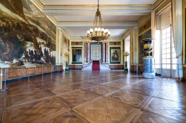 Oda ahşap zeminli ve duvarlarında resimler olan geniş bir alan gösteriyor. Avizeler ve büyük bir merdiven var..