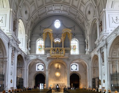 İnsanlar Münih 'teki St. Michael Kilisesi' nde toplanıyor. İç kısımda renkli tasarımları olan bir organ ve büyük pencereler var..