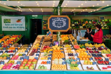 Ziyaretçiler Viktualienmarkt, Münih 'teki bir pazarda taze meyve ve sebzelere bakıyorlar. Pazar çeşitli ürünler ve yerel ürünler sunmaktadır..
