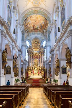 Ziyaretçiler Münih 'teki Aziz Peter kilisesini keşfediyorlar. Parlak ışık pencerelerden süzülüyor. Sunak ve süslü detaylar dikkat çeker..