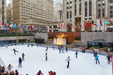 İnsanlar Manhattan 'daki Rockefeller Center' da yoğun bir kış gününde buz pateni pistinde kayıyorlar. Aileler izlerken bayraklar dalgalanıyor ve katılıyorlar.