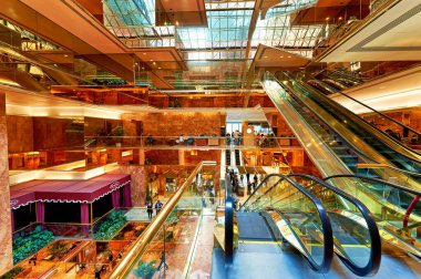 Yürüyen merdivenler inip çıkarken insanlar Trump Kulesi 'nin avlusunda yürüyor ve etkileşime giriyorlar. Boşlukta uzun duvarlar ve geniş pencereler var..