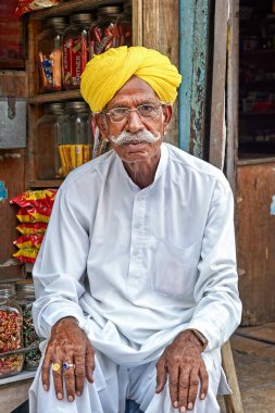 Jaisalmer 'de bir markette kıdemli bir adam oturuyor. Geleneksel kıyafetler ve sarı bir türban giyiyor. Etrafındaki işlek sokağı izliyor..