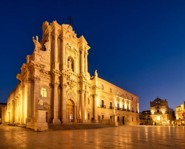 İnsanlar geceleri Ortygia Adası 'ndaki Katedral' in yanında yürürler. Bina, karanlık gökyüzüne karşı parlak ışıklar gösteriyor..