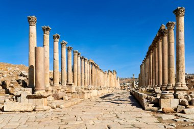Ziyaretçiler Ürdün 'ün Gerasa kentindeki sütunlu cadde boyunca yürüyorlar. Taş sütunlar açık mavi bir gökyüzünün altında dimdik duruyorlar..