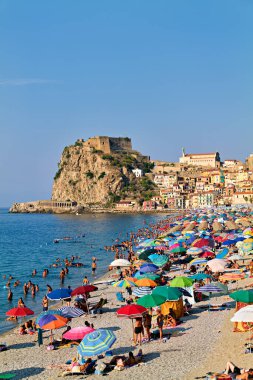 İnsanlar yazın Scilla, Calabria 'daki Marina Grande plajında renkli şemsiyeler ve arka planda Ruffo Kalesi ile eğleniyorlar..