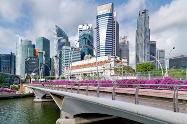 İnsanlar Singapur 'un mali bölgesinde bir köprünün yanından geçiyorlar. Arka planda yüksek binalar ve açık gökyüzü görülüyor.
