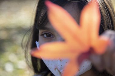 Portakallı sonbahar yaprağı tutan morina yüzünden maske takan genç bir kız. Eş zamanlı yatay görüntü. Sonbahar konsepti.