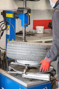 Gri üniformalı ve kırmızı eldivenli tamirci, atölyede lastiği değiştirmek için lastiği kenardan çıkarıyor..