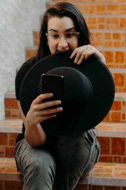 Cep telefonu kullanan, gözlüklü, siyah şapkalı bir kadının portresi..