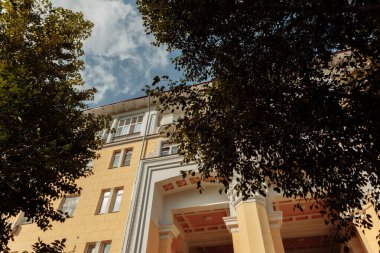 Güneş tarafından aydınlatılan sarı bir bina ağaçlardan dışarı bakıyor. Bina Stalinist İmparatorluk tarzında inşa edildi. Yüksek kalite fotoğraf