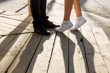 Öpüşen çift. Beyaz spor ayakkabılı kadınların bacakları parmaklarının ucunda. Siyah çizme giymiş erkek ayakları yakınlarda duruyor..