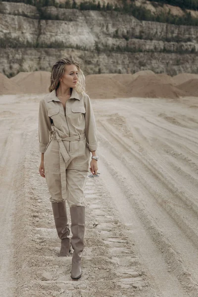 Sarışın bir kadın kumda yürüyor ve yan tarafa bakıyor. Kız çalışan bir tulum giyiyor. Kızın elinde bardaklar var. Yüksek kalite fotoğraf