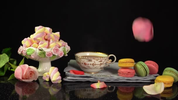 Composizione con una tazza di tè, amaretti, marshmallow e petali di rosa cadenti — Video Stock