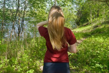 Uzun saçlı, arka görüşlü bir kız. Kırmızı tişörtlü bir kadın doğada duruyor ve kahverengi saçlarını düzeltiyor.