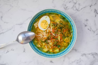 Mavi tabakta Udon erişte çorbası ve yumurta, patates ve taze bitkilerle metal kaşık.