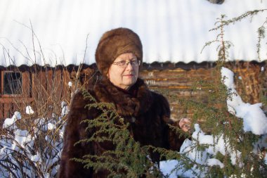 Yetişkin bir kadın, kışın sokakta duruyor. Şapkası ve kürkünden yapılmış kürkü var. Yüzü, akşam güneşiyle aydınlanıyor.