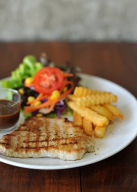 Bir tabak ızgara biftek, patates kızartması ve ahşap arka planda salata.