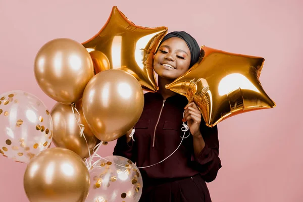 Christmas celebration. African american holiday. Attractive girl with golden helium balloons isolated peach background. Young black woman weared traditional dress and scarf.
