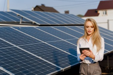Yatırımcı kadın elinde tabletle mavi güneş panellerinin yanında duruyor. Kız resmi beyaz gömlek giyiyordu. Ev için bedava elektrik. Gezegenin sürdürülebilirliği. Yeşil enerji.