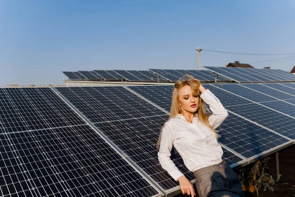 Woman leans on solar panels. Blonde dressed white formal shirt on the power plant. Free electricity for home. Green energy. Solar cells power plant business.