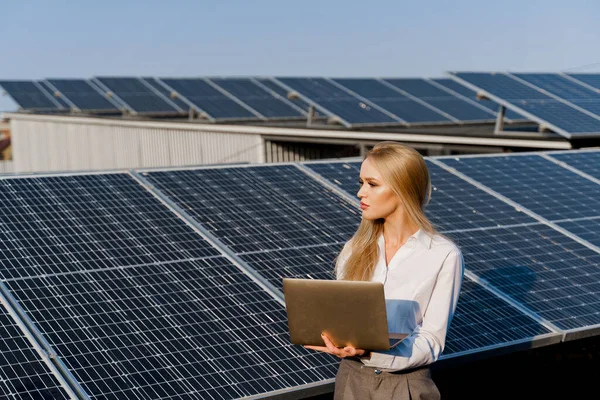 Investor woman stand with laptop near blue solar panels row on the ground Girl weared formal white shirt. Free electricity for home. Sustainability of planet. Green energy.