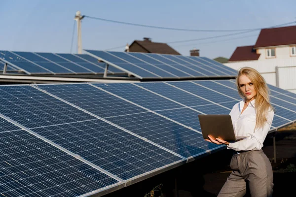 Investor and solar panels. Blonde woman with laptop near blue solar panels row on the ground Girl weared formal white shirt. Free electricity for home. Sustainability of planet. Green energy.