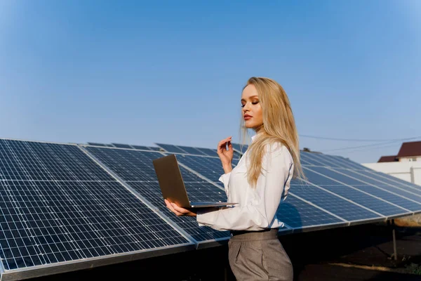 Investor and solar panels. Blonde woman with laptop near blue solar panels row on the ground Girl weared formal white shirt. Free electricity for home. Sustainability of planet. Green energy.