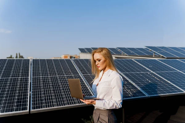 Investor and solar panels. Blonde woman with laptop near blue solar panels row on the ground Girl weared formal white shirt. Free electricity for home. Sustainability of planet. Green energy.
