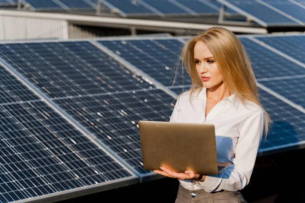 Investor woman stand with laptop near blue solar panels row on the ground Girl weared formal white shirt. Free electricity for home. Sustainability of planet. Green energy.
