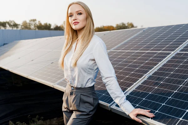Blonde model with solar panels stands in row on the ground. Girl dressed white formal shirt smiles on the power plant. Free electricity for home. Green energy. Solar cells power plant business.