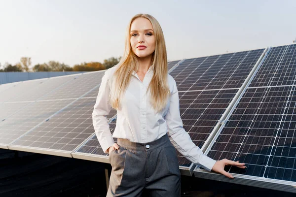Blonde model with solar panels stands in row on the ground. Girl dressed white formal shirt smiles on the power plant. Free electricity for home. Green energy. Solar cells power plant business.