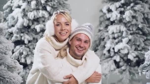 幸せな若いカップルの抱擁と笑顔と雪は冬の日に新年のお祝いの前夜にクリスマスツリーの近くに落ちます。笑顔の男と女は白い恋人を身に着けていた愛し合う — ストック動画