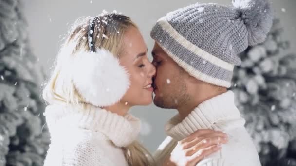 雪とキスだ。冬の日のお祝いの前夜に、幸せな若いカップルがクリスマスツリーの近くで抱擁とキスをします。笑顔の男と女は白い恋人を身に着けていた愛し合う — ストック動画