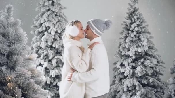 Spadający śnieg i buziaki. Szczęśliwa młoda para przytula się i całuje w pobliżu choinek w przeddzień Nowego Roku w zimowy dzień. Uśmiechnięty mężczyzna i kobieta nosili białe pulowery kochają się — Wideo stockowe