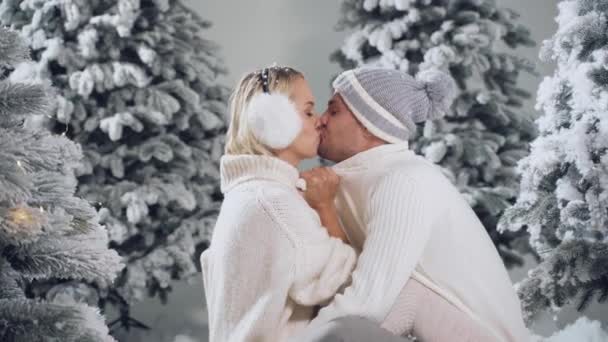 新年爱情故事。情侣的亲吻和拥抱。人们穿着毛皮耳机、帽子、白色毛衣。冬日快乐的年轻夫妇在圣诞树旁拥抱亲吻. — 图库视频影像