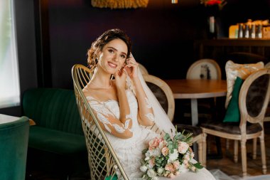 Düğün buketi olan gelin gülümsüyor, kameraya bakıyor ve yüzüne dokunuyor. Sosyal ağlar için çekici bir kız portresi. Lüks restoranda gelinlikli bir kız.