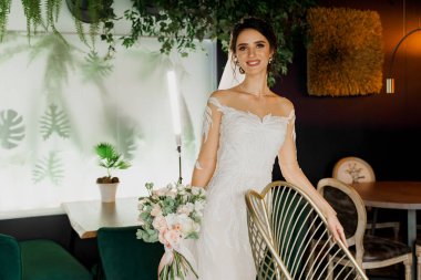 Düğün buketi olan gelin gülümsüyor, kameraya bakıyor ve yüzüne dokunuyor. Sosyal ağlar için çekici bir kız portresi. Lüks restoranda gelinlikli bir kız.