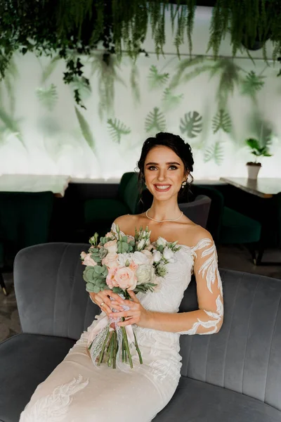 Bride with wedding bouquet smiles, looks in camera and touches her face. Attractive girl portrait for social networks. Girl in wedding dress in luxury restaurant