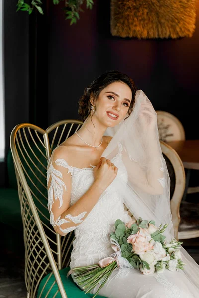 Bride with wedding bouquet smiles, looks in camera and touches her face. Attractive girl portrait for social networks. Girl in wedding dress in luxury restaurant