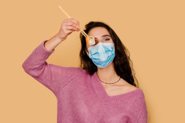 Suşi maskeli kız sarı arka planda izole edilmiş. Genç bir kadın, Philadelphia usulü yemek çubuklarıyla suşi tutuyor. Coronavirus covid-19 karantinasında suşi teslimatı yapılacak.