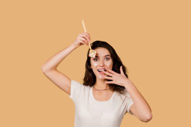 Suşili mutlu kız sarı arka planda izole edilmiş. Genç kız elinde yemek çubuklarıyla suşi tutuyor, gülümsüyor, eğleniyor ve bir dilim poğaça yemek istiyor. Suşi teslimatı için yönlendir.