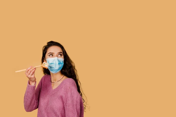 戴着医用面罩的寿司女孩被隔离在黄色背景下 年轻的女人拿着寿司用的是筷子片的胶卷 在检疫Coronavirus Covid 19时作寿司派递广告 — 图库照片