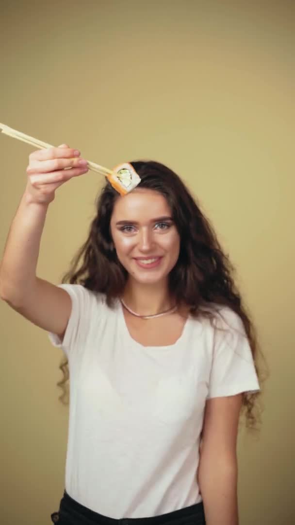 Happy girl giving sushi to you isolated on yellow background vertikális videó. Fiatal lány tartja sushi evőpálcikával, mosolyog, szórakozik, és akar enni egy darab tekercs. Hirdetés sushi szállításra — Stock videók