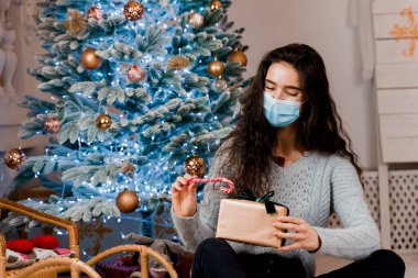 Noel hediyesi ve şekerle birlikte tıbbi maskeli bir kız. Kendini izole etme üzerine yeni yılı kutluyoruz. Salgın koronavirüs covid-19 konsepti. Karantina döneminde reklam indirimi