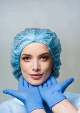 Beyaz arka planda estetik cerraha danışmanlık. Kozmetik yenileme yüz tedavisi. Mavi eldivenli doktor kız yüzüne dokunuyor. Hasta dudakları ve çene enjeksiyonlarını soruyor.