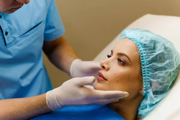 Estetik cerraha danışmanlık. Kozmetik yenileme yüz tedavisi. Maskeli doktor kız hastaya dudaklarına, çenesine ve alnına yapılan iğneleri soruyor. Kırışıklık giderici